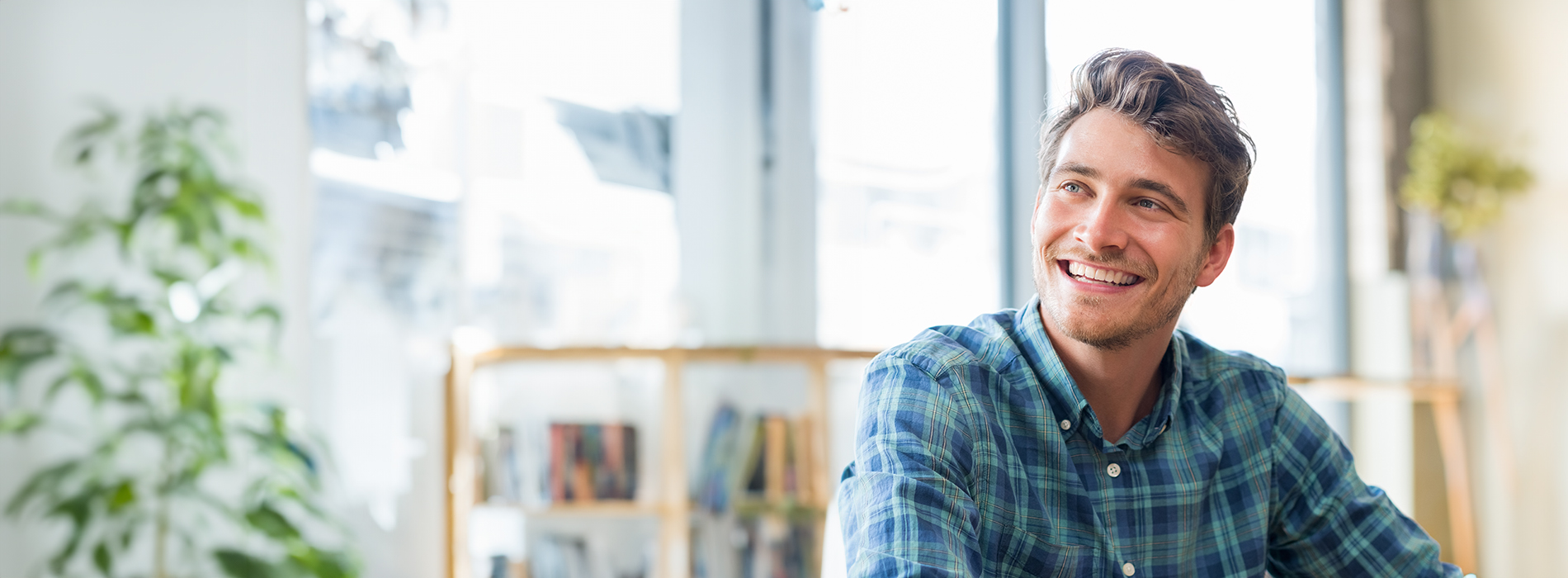 A man with a beard and mustache is seated at a desk with a green checkered shirt, smiling towards the camera.