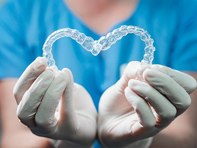 A hand holding a transparent dental retainer shaped like a heart, with a blue surgical glove in the background.