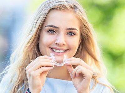 A young woman with blonde hair is smiling and holding up a clear dental retainer, showcasing her smile.