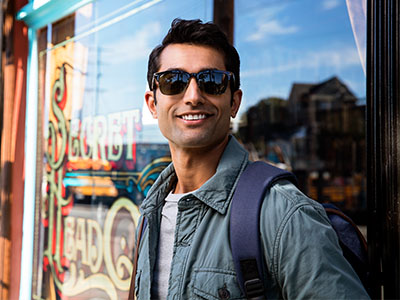 A man in sunglasses and a backpack is standing outside a storefront, smiling towards the camera.