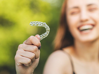 The image shows a young woman smiling and holding up a clear plastic dental retainer.