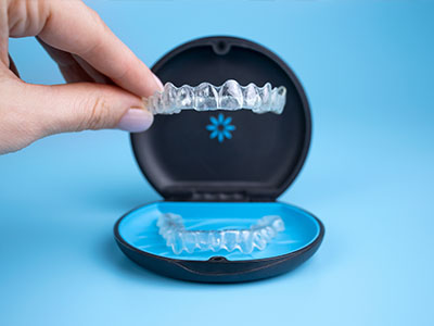 The image shows a person s hand holding an open case with a clear plastic dental retainer inside, which is placed on top of a blue box.