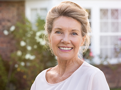 The image shows a smiling older woman with blonde hair, wearing a white top and standing in front of a house.