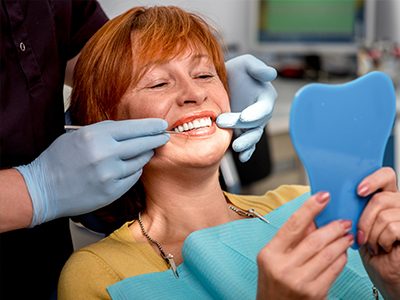 A woman in a dental chair receiving a teeth cleaning, with a dentist performing the procedure and holding up a blue dental model.