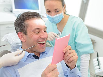 The image features a man sitting in a dental chair, smiling at the camera while holding up a pink card with both hands. Behind him, a female dental professional is standing and appears to be showing him something on a tablet or screen. They are both in a dental office setting, with medical equipment visible around them.