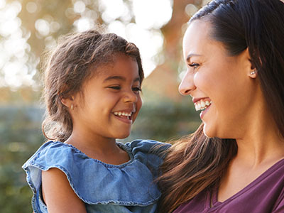 The image features a woman and a young child, both smiling at the camera. The woman is wearing a sleeveless top and has long hair, while the child appears to be a toddler with short hair, dressed in a light-colored shirt. They are standing outdoors during daylight, with greenery visible behind them.