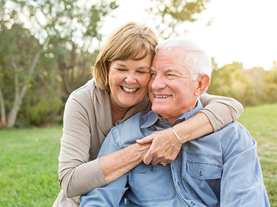 The image shows a man and woman, both elderly, embracing each other in a warm pose. They are outdoors during daylight, with the man wearing a blue shirt and the woman in a white top.