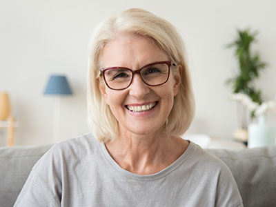 A smiling woman with short blonde hair, wearing glasses and a light-colored top, seated indoors against a neutral background.