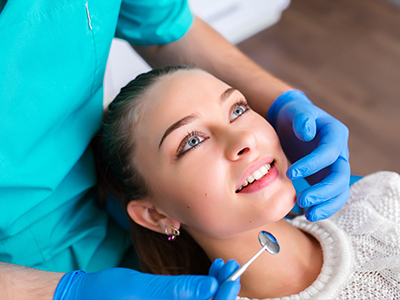 In the image, a young woman is seated in a dental chair with her eyes closed. A dentist is adjusting a clear dental appliance on her teeth while she smiles gently.