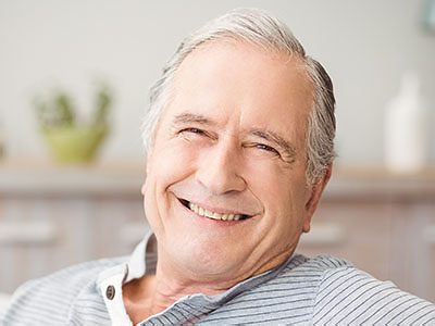 A smiling older man with gray hair, wearing a blue shirt and glasses, sitting in a chair.