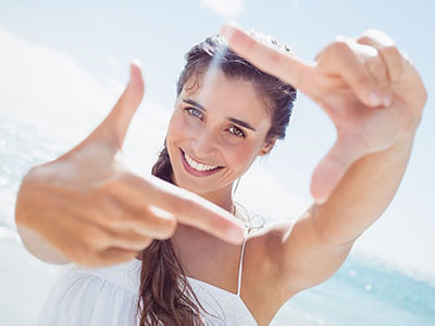 A woman with long hair is smiling and holding up a hand with fingers spread wide, creating a frame with her hand in front of her face.